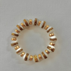 Puede incluir: Un brazalete de cuentas blancas y doradas con un patrón a rayas. El brazalete está hecho de cuentas redondas con una raya dorada alrededor del medio.