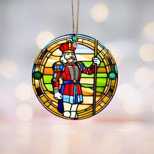 May include: A round, colorful stained glass ornament featuring a Nutcracker soldier holding a spear. The ornament has a gold frame and a green, yellow, and orange background.