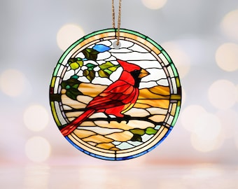 May include: A round, stained-glass ornament featuring a vibrant red cardinal perched on a branch with green leaves. The bird is set against a backdrop of yellow and tan hues, framed by a circular border with a gold string for hanging.