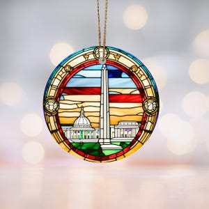 Może przedstawiać: Okrągła szklana ozdoba z witrażowym wzorem przedstawiającym Pomnik Waszyngtona i Kapitol. Ozdoba jest otoczona czerwoną, białą i niebieską ramką.