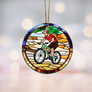 May include: A round stained glass ornament featuring a person riding a mountain bike on a dirt path. The ornament is decorated with a colorful landscape and a blue, red, and yellow border.