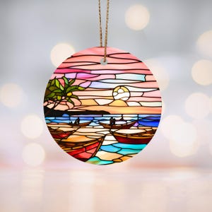 Peut inclure: Une décoration de Noël en verre teinté représentant un coucher de soleil sur une plage avec des palmiers et des bateaux. La décoration est ronde et présente une palette de couleurs rose, orange et bleue.