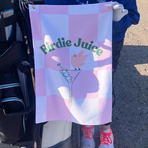 May include: A pink and white checkered golf towel with the text "Birdie Juice" in green letters. A pink bird with green accents is perched on a martini glass with green olives.