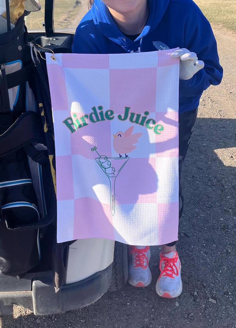 May include: A pink and white checkered golf towel with the text "Birdie Juice" in green letters. A pink bird with green accents is perched on a martini glass with green olives.