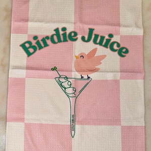 May include: Pink and white checkered golf towel with the text "Birdie Juice" in green letters. A pink bird is perched on a martini glass with green olives. The text "Golf Girllies" is written on the stem of the glass.