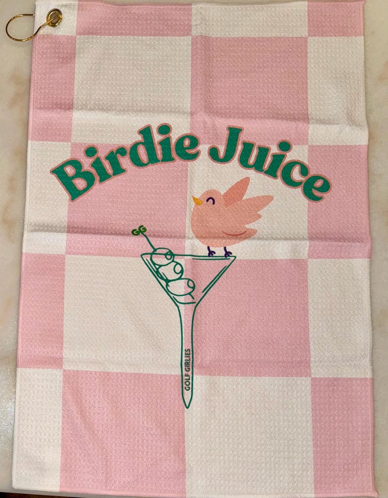 May include: Pink and white checkered golf towel with the text "Birdie Juice" in green letters. A pink bird is perched on a martini glass with green olives. The text "Golf Girllies" is written on the stem of the glass.