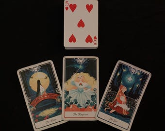 May include: A collection of tarot cards and a playing card with a black background. The tarot cards feature illustrations of The Moon, The Magician, and The Star. The playing card shows the number 5 and five red hearts.