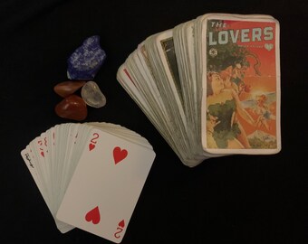 May include: A deck of tarot cards with the title "The Lovers" on the card face, featuring an illustration of two figures. A separate deck of playing cards with red hearts is also visible, along with several polished gemstones.