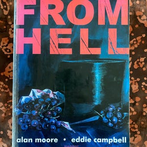 Op de afbeelding: Boekomslag met de titel "FROM HELL" in grote, roze, verweerde letters. De omslag heeft een donkerblauwe achtergrond met een illustratie van een hoge hoed en druiven. De namen van de auteurs, Alan Moore en Eddie Campbell, staan onderaan.