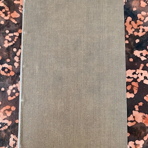 Op de afbeelding: Een gesloten rechthoekig boek met een olijfgroene, getextureerde kaft. De achtergrond heeft een abstract patroon in oranje en bruine tinten. Het boek lijkt in goede staat te zijn. De kaft heeft een subtiel, geweven patroon.