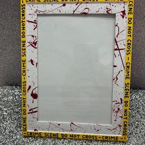 May include: A rectangular photo frame with a white background and red splatter paint design. The frame is bordered with yellow tape that reads "CRIME SCENE DO NOT CROSS - CRIME" in black lettering. The frame is designed to hold a 5x7 inch photo.