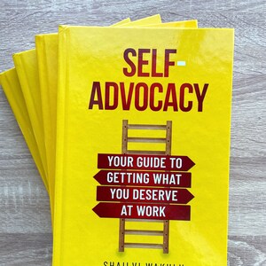 May include: A yellow book titled "Self-Advocacy: Your Guide to Getting What You Deserve at Work" by Shailvi Wakhlu. The book cover features a wooden ladder with red signs that read "Your Guide to Getting What You Deserve at Work."