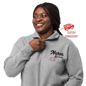 May include: A grey quarter-zip sweatshirt featuring the text "Nurse Cindy" and a pink stethoscope design. The sweatshirt has a high collar and a zip closure. The image also includes the text "Custom Embroidery!"