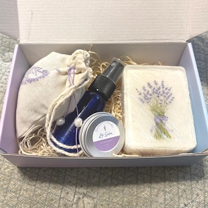 May include: A gift box filled with lavender-themed bath and body products. The box contains a small bag of lavender buds, a bottle of lavender spray, a small tin of lavender lip balm, and a bar of lavender soap.