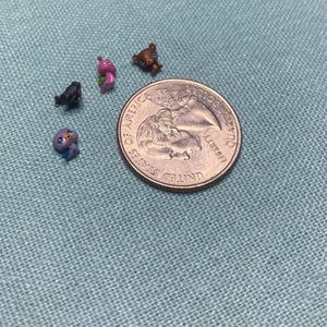 May include: Four miniature animal figurines in purple, black, pink, and brown, positioned next to a United States quarter coin for scale. The tiny figures feature large eyes and distinct animal shapes, resting on a textured light blue surface.