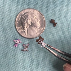 Puede incluir: Cuatro figuras de animales en miniatura que incluyen una criatura morada, un gato siamés, un perro marrón y blanco sostenido por pinzas de metal y una pequeña mancha marrón. Se incluye una moneda de 25 centavos de EE. UU. para escala.