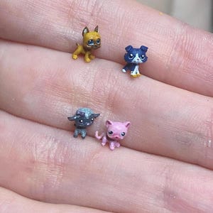 May include: Four miniature animal figurines including a yellow dog, a dark blue and white dog, a dark gray creature with a light blue head, and a small pink cat. These tiny, detailed collectibles are displayed together, showcasing distinct colors.