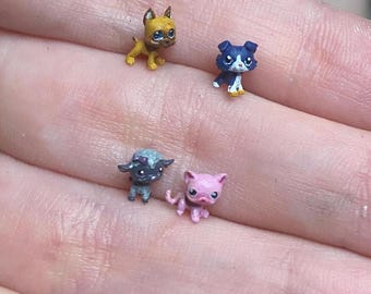 May include: Four miniature animal figurines including a yellow dog, a dark blue and white dog, a dark gray creature with a light blue head, and a small pink cat. These tiny, detailed collectibles are displayed together, showcasing distinct colors.