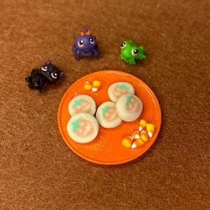 Op de afbeelding: Een oranje miniatuurbordje met vijf ronde koekjes met oranje pompoenontwerpen en verschillende kleine stukjes suikermaïs, vergezeld van drie kleine plastic dierenfiguurtjes: een zwarte kat, een paars wezen en een groene kikker.