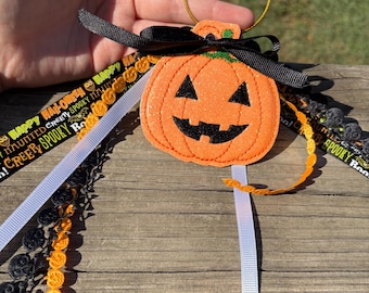 May include: Orange pumpkin-shaped Halloween keychain with a black smiling face, black bow, and decorative ribbons. The ribbons include the text "Happy Halloween" and "Creepy Spooky Boo". The keychain has a gold-colored ring.