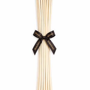 May include: A bundle of long, slender, light beige reed diffuser sticks tied together at the center with a black satin ribbon. The ribbon features the gold-colored text MooseScents printed repeatedly along its length in a decorative font.