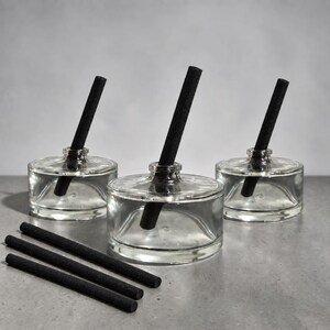 May include: Three clear glass circular vessels with short necks, each containing a single black cylindrical reed. Three additional black cylindrical reeds are positioned horizontally in the foreground, separate from the glass containers.