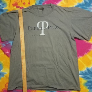 Puede incluir: Camiseta gris con el texto blanco "Pepe Le Pew" y "Skunkwear" debajo. La camiseta está colocada plana, mostrando el diseño frontal. Una regla de madera se coloca verticalmente sobre la camiseta.