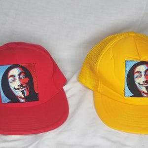 Puede incluir: Dos gorras de béisbol con un gráfico de una persona que lleva una máscara de Guy Fawkes. Una gorra es roja y la otra es amarilla.