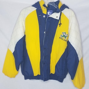Puede incluir: Una chaqueta vintage Starter azul, amarilla y blanca con un logotipo de Notre Dame y las palabras "Notre Dame" bordadas en la parte delantera. La chaqueta tiene una cremallera y una capucha.