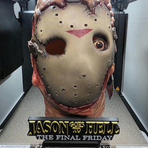 May include: A detailed replica mask featuring a weathered, tan hockey-style faceplate with a red triangle accent, mounted on a textured, flesh-toned base. A black display plaque in front reads JASON GOES TO HELL THE FINAL FRIDAY in yellow lettering.