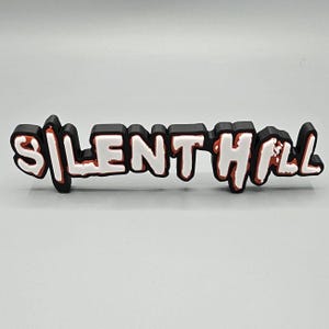 May include: A black 3D printed sign with the words "SILENT HILL" in white letters with a red outline.