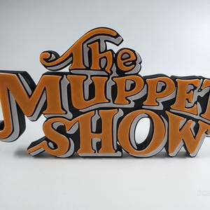 May include: A three-dimensional sign with the words "The Muppet Show" in orange, black, and white. The sign has a retro design with a playful font, ideal for fans of the classic television show.