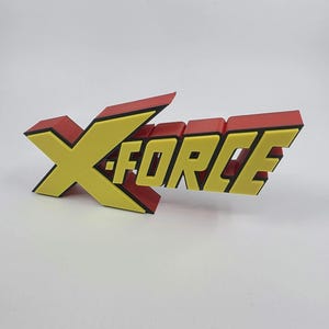 Könnte beinhalten: Ein 3D-gedrucktes X-Force-Logo in Gelb und Rot. Das "X" ist gelb mit roter Umrandung, und der Text "FORCE" ist gelb mit schwarzer Umrandung. Das Logo wird auf einer weißen Oberfläche angezeigt.