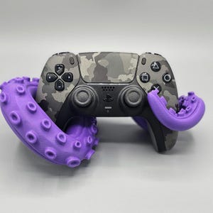 May include: A camouflage-patterned video game controller is held by a purple tentacle-shaped stand. The controller has black buttons and a touchpad. The stand has suction cup details.