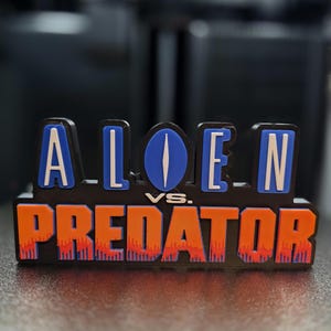 以下が含まれることがあります： 「ALIEN vs. PREDATOR」と書かれた黒いサイン。 「ALIEN」の文字は青と白で、中央に青い楕円があります。 「PREDATOR」はオレンジ色で赤いアクセントが付いています。 ポップカルチャーアイテム。