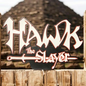 Könnte beinhalten: Dekoratives Schild mit dem Schriftzug "HAWK The SLAYER" in einer stilisierten Schriftart. Die Buchstaben sind weiß mit roten und schwarzen Akzenten. Das Schild ist an einem Holzzaun befestigt, mit einer Pyramide im Hintergrund.