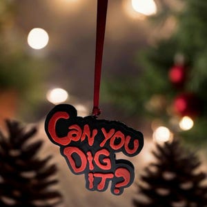 Peut inclure: Une décoration de Noël noire avec une inscription rouge qui dit "Can You Dig It?" suspendue à un ruban rouge.