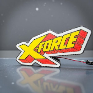 以下が含まれることがあります： 黄色い「X」と赤い「FORCE」の文字が入ったカラフルなX-FORCEロゴ。ロゴは白い縁取りがあり、複数の色のストランドを持つワイヤーに接続されています。背景はぼやけたグレーです。