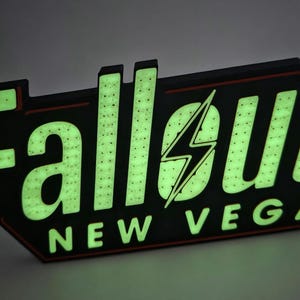 May include: A glowing sign with the words "Fallout New Vegas" in green, with a black and red border. The sign features a lightning bolt graphic and is designed to resemble a retro motel sign.