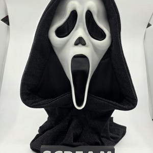 Op de afbeelding: Een Scream-masker met een wit gezicht en zwarte oogkassen, mond en kap. Het masker wordt gepresenteerd op een zwarte standaard met het woord "SCREAM" in witte letters.