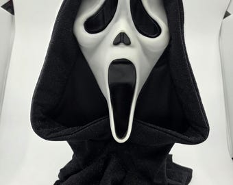 May include: A Scream mask with a white face and black eye sockets, mouth, and hood. The mask is displayed on a black stand with the word "SCREAM" in white letters.