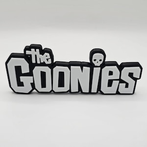 May include: Black and white 3D printed sign with the text "The Goonies" in bold white letters with a black skull above the word "Goonies".