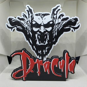 May include: A black and white Dracula-themed decorative piece featuring a stylized image of a vampire's face. The word "Dracula" is displayed in red, with a black outline, on a black base. The design is 3D.