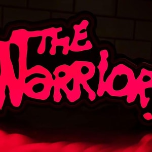 May include: A neon sign with the words "The Warriors" in a pink, graffiti-style font on a black background.