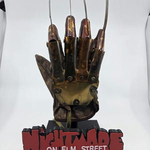 May include: A replica glove with metal blades, inspired by the horror film "A Nightmare on Elm Street." The glove is brown and metallic, with a leather-like texture. The base reads "A Nightmare on Elm Street" in red letters.