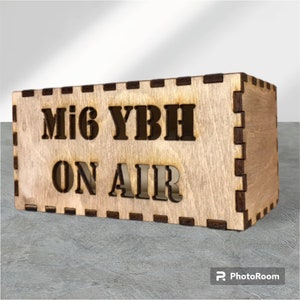 Puede incluir: Caja rectangular de madera clara con las letras "Mi6 YBH ON AIR" recortadas. Los bordes son más oscuros. El objeto está colocado sobre una superficie gris.