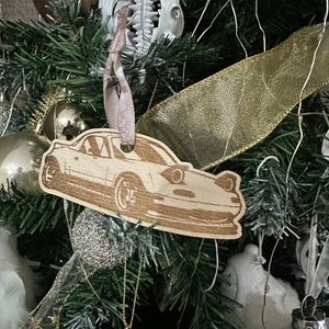 May include: A wooden ornament shaped like a car, hanging on a Christmas tree with gold and white decorations. The ornament is a light brown colour and has a detailed design.