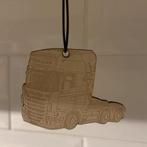 May include: A wooden truck-shaped ornament. The light brown ornament features etched details of a lorry, including the words "TOP LINE" and "SCANIA". It hangs from a black string.