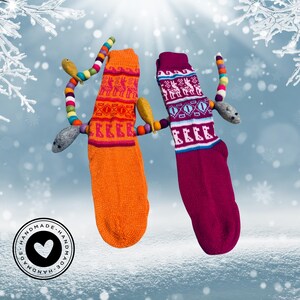May include: Two colorful knitted socks with a whimsical felt snake design. The socks are orange and pink with a geometric pattern. The snake is made of felt and has a variety of colors, including green, blue, yellow, and gray. The socks are handmade and have a "Handmade" stamp on the bottom.