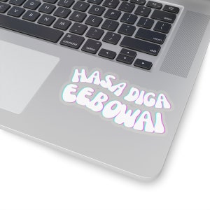 May include: A white sticker with the text "HASA DIGA EEBOWAI" in a retro font, outlined in blue and pink, is affixed to a laptop. The laptop is silver and has a black keyboard.